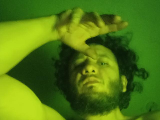 DavydWolfy69, avec ses cheveux noirs bouclés, sa barbe et sa peau légèrement verdâtre, couvrant son œil droit avec sa main gauche, torse nu, regardant vers le haut avec une expression légèrement confuse..