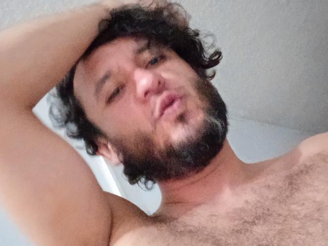 DavydWolfy69 tiene una barba negra y rizada, cabello oscuro y ondulado, y un pecho cubierto de vello, mirando hacia arriba con la boca ligeramente abierta, levantando un brazo detrás de la cabeza..