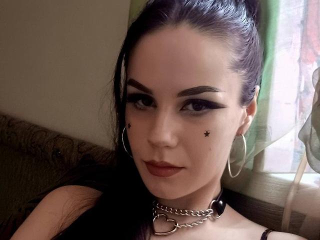Cassie Evans, avec des cheveux noirs attachés en queue de cheval, du crayon noir pour les yeux, des tatouages en forme d'étoiles près de ses yeux, du rouge à lèvres rouge, des boucles d'oreilles cerceaux, un collier en argent, assise sur un canapé à motifs verts..