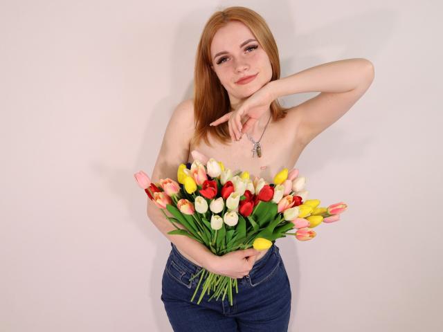 MargaritaKis, oben ohne, mit roten, gelben und rosa Tulpen, trägt blaue Jeans, hält einen Blumenstrauß, rechter Arm erhoben, linker Arm über der Brust, leichtes Lächeln, Halskette, helle Haut, rotes Haar..