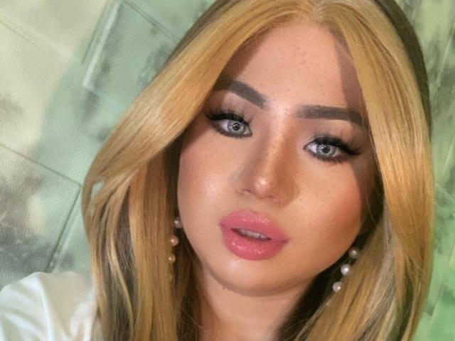 NaughtyRosecum has blonde hair, blue eyes, thick eyebrows, and pink glossy lips. She's wearing pearl earrings and has a slightly open, seductive mouth. Her skin is fair and freckled.