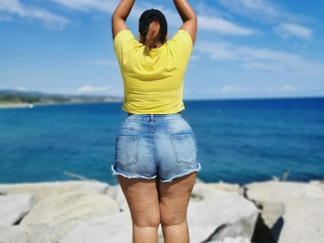LouiseSexBigBoobs with her back to the camera, arms raised, wearing a yellow shirt and frayed blue denim shorts, standing on rocky beach with blue ocean and sky.