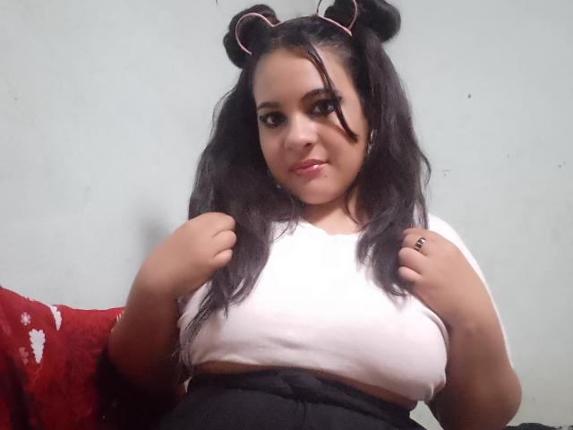 Merilylu with black hair in bear ear headbands, white crop top, showing her big tits, hands near her chest, slightly pouty expression, red pillow in background.