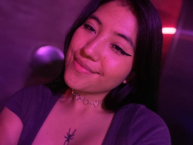 Elizaa, com cabelo preto, sorriso adorável, pele rosada, delineador, colar e uma tatuagem de aranha no peito, vestindo uma blusa roxa, em iluminação roxo-avermelhada..