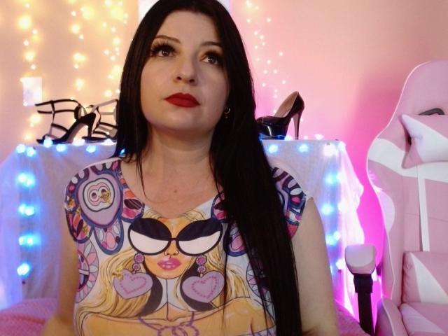 NatalyHami with long black hair, red lipstick, and a colorful graphic shirt featuring a face with sunglasses and hearts. She's sitting in a pink gaming chair.