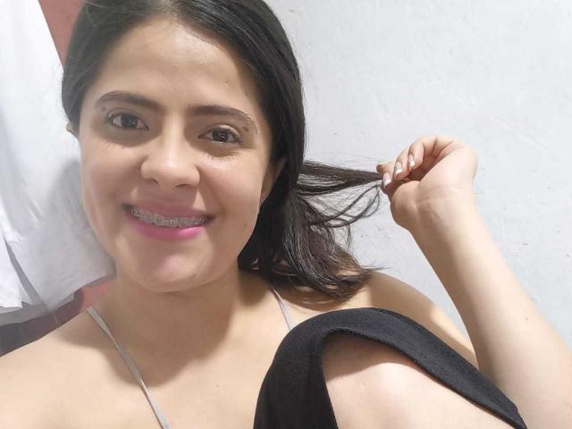 StephanyGrecie with dark hair, smiling, holding a hair strand, wearing a black strap, grey halter, showing metal braces, pink lipstick, and a cute face, against a white wall.