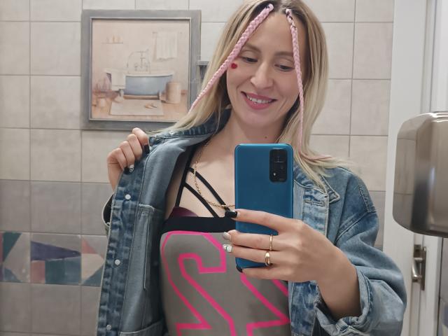 Saveta, avec ses cheveux tressés roses, sa veste en jean et son t-shirt graphique rose, montre ses seins dans un haut noir à bretelles fines, souriant dans un selfie pris devant le miroir de la salle de bain, tenant un téléphone bleu..