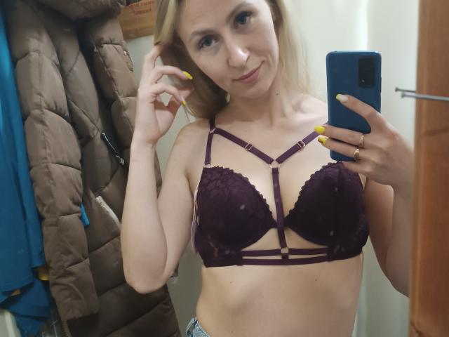 Saveta with small tits in a purple lace bra, holding a blue phone, yellow nails, smiling, blonde hair, in a mirror selfie, wearing blue jeans, in a bathroom.