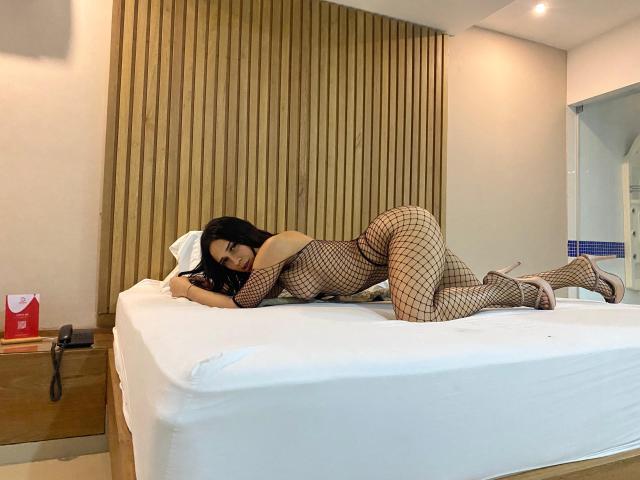 SamysPresleyTs is lying on a bed, wearing a fishnet bodysuit, showing her ass, with black hair, and pink high heels, in a bedroom, looking at the camera.