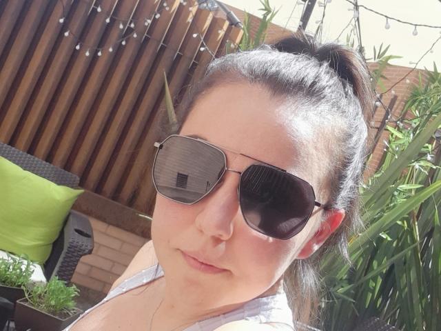 AdelaShery with brown hair in a ponytail, wearing aviator sunglasses, and a white top, has a slightly chubby face, light skin, and a neutral expression in a sunny outdoor setting.