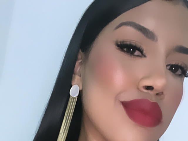 StephanieDavies has dark hair, thick eyebrows, long eyelashes, red lipstick, and gold earrings with a white circular top. She's smiling with a slightly flushed face.