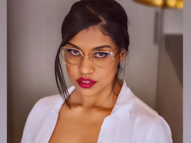 StephanieDavies has dark hair, wears glasses, and red lipstick, with a white shirt exposing her tits. She's looking at the camera with a serious expression.