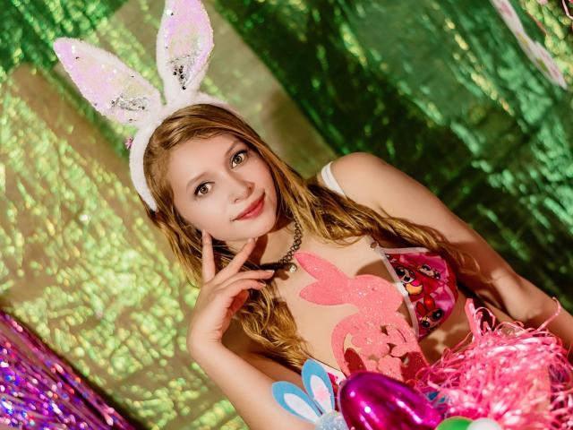 KeiliSexy with blonde hair, wearing white bunny ears, pink glittery heart chest piece, and pink fluffy dress, smiling, pointing to her lips, holding pink and blue accessories.