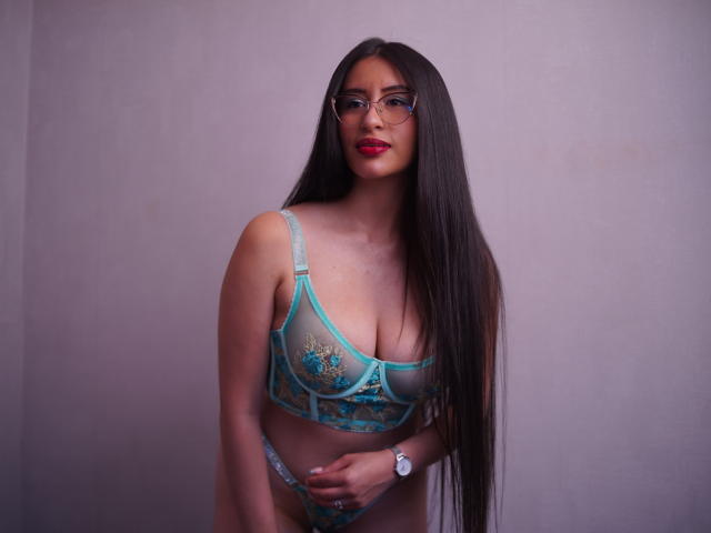 CassieSmith with long dark hair, glasses, and red lipstick, wearing a turquoise lace bra and matching panties, showing her tits and holding her pussy. Wearing a white watch.