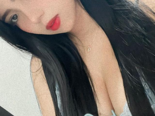 Bella Dawson, met lang zwart haar, rode lippenstift en een gouden ketting, laat haar decolleté zien in een grijze top en kijkt met verleidelijke ogen naar beneden. Haar borsten zijn zichtbaar..