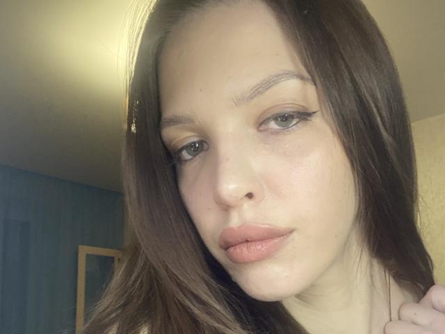 KateMariett has light brown hair, green eyes with black eyeliner, and pink lips. Her fair skin is slightly shiny with a small nose. She's looking at the camera.