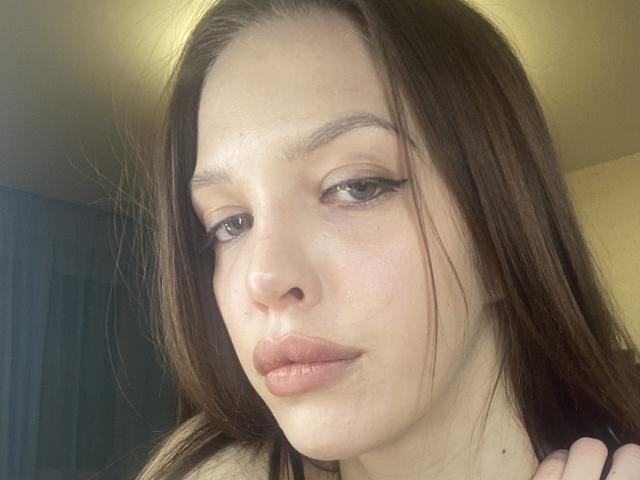 KateMariett has brown hair, light skin, and full pink lips. She has thick eyebrows, long eyelashes, and a slight pout. Her hand is near her chin.
