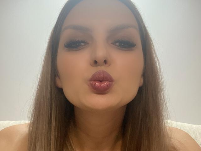 DouceAmelia's pouty lips are puckered up like a small, pink, shiny butt. She's got long brown hair, thick eyelashes, and a neutral expression. Light skin, straight hair.