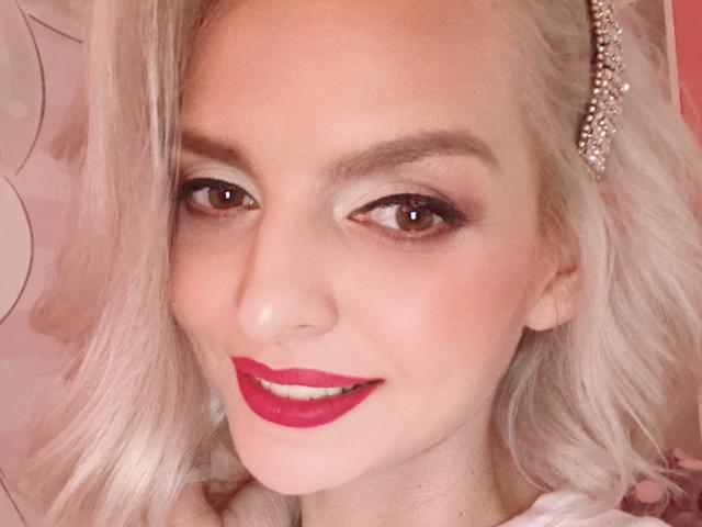 DouceAmelia has platinum blonde hair, red lipstick, and a pearl hairpin. Her makeup is smoky, and she's smiling with her brown eyes looking at the camera.
