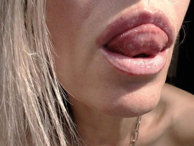 VickySexyFr's pink tongue is fully out, touching her glossy, plump lips. She has light blonde hair, fair skin, and a slight shadow on her lower chin.