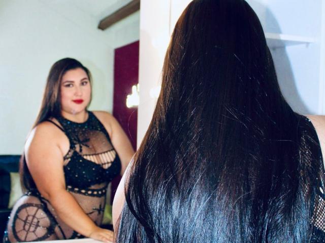 LarysaTayloor, com longos cabelos pretos, olhando para o espelho, vestindo lingerie de malha preta com recortes, mostrando seu grande bumbum e seios, em pé em um quarto com uma porta vermelha..