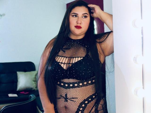 LarysaTayloor with long black hair, wearing a black mesh lingerie dress, revealing her big tits and black bra, standing by a white wall, hand in hair.