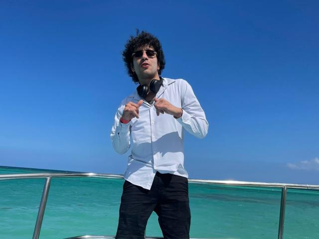 Damian Delatorre, avec ses cheveux noirs bouclés, portant des lunettes de soleil, une chemise blanche, un pantalon noir, tenant des écouteurs noirs, debout sur une rambarde en métal, ciel bleu, océan turquoise clair, ensoleillé..