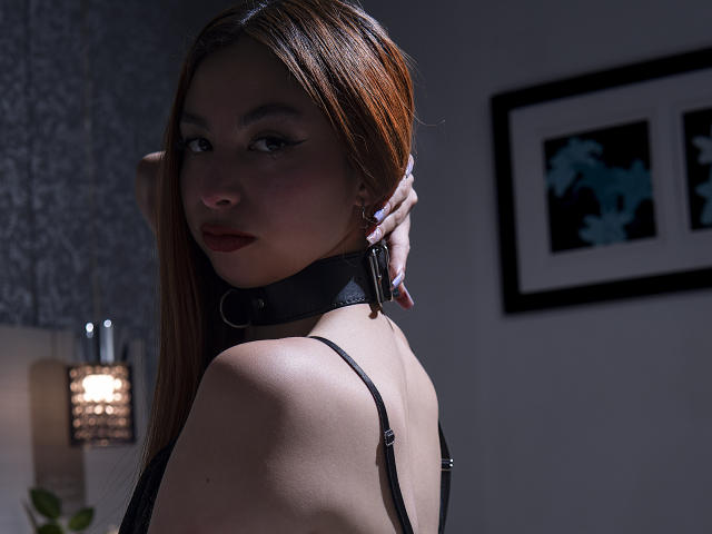 CamilleRios met bruin haar, draagt een zwarte leren halsband, kijkt terug naar de camera, zwarte jurk met bandjes, zachte belichting, hand op de nek, subtiele make-up, donkere nagellak..