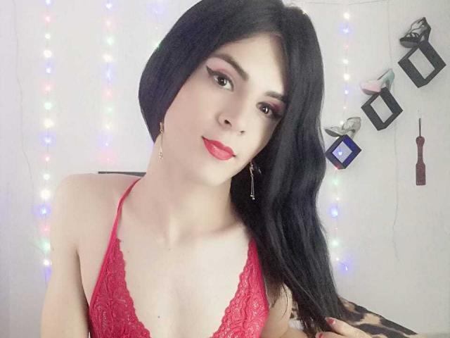 DanielaMonty with long black hair, red lace halter top, pink eyeshadow, red lipstick, and small gold earrings, smiling gently. Boobs visible, soft light, relaxed expression.