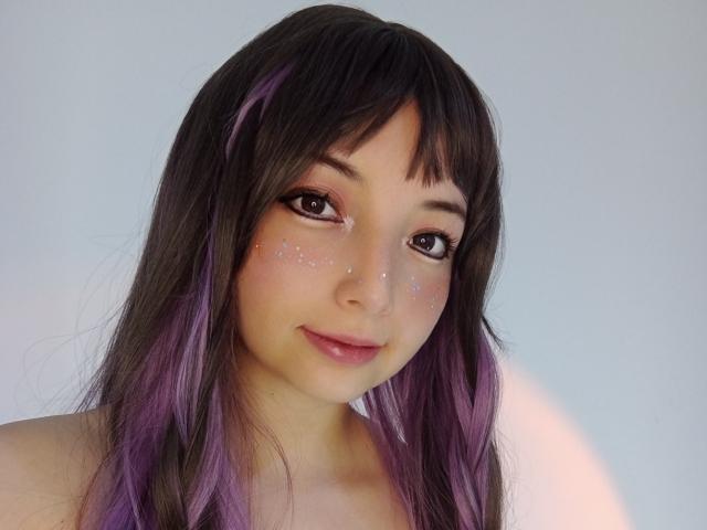 BarbaraJonesX has long, dark brown hair with purple tips, sparkling eyes, and a cute smile, wearing subtle glitter on her cheeks. She's topless, with a soft, natural look.