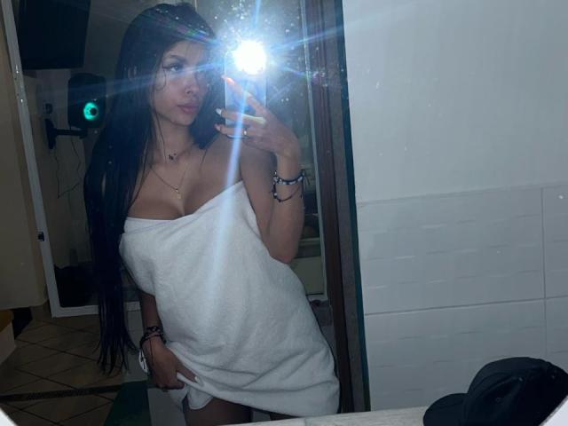 NatyJanssen with long dark hair, white towel covering her tits and ass, taking a mirror selfie with a bright flash, wearing bracelets, in a dimly lit bathroom.