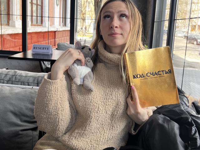 Saveta, com cabelo loiro, vestindo um suéter bege felpudo, segurando um livro dourado e um pequeno brinquedo de pelúcia, sentada em um café. Seus seios são pequenos e ela é fofa..