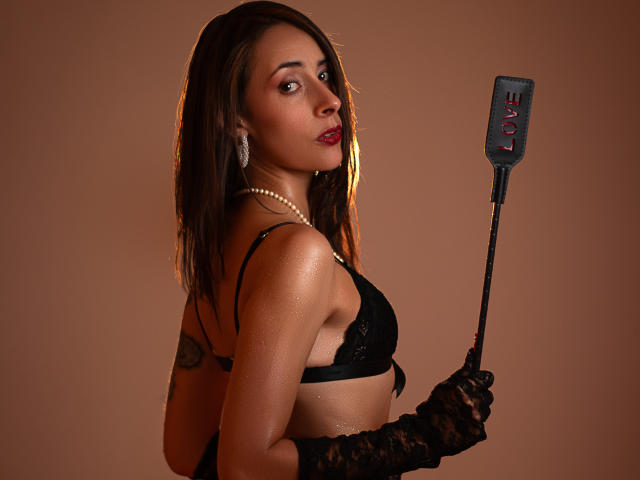 ScarletKings with glistening skin, black lace gloves, and pearl necklace, holding a black flogger, wearing a black bra, and red lipstick, looking over her shoulder.