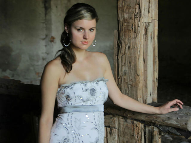 Darcie-hot with pale skin, wearing a strapless white dress with silver patterns, standing by a wooden pillar, has brown hair in a ponytail, wearing large hoop earrings.