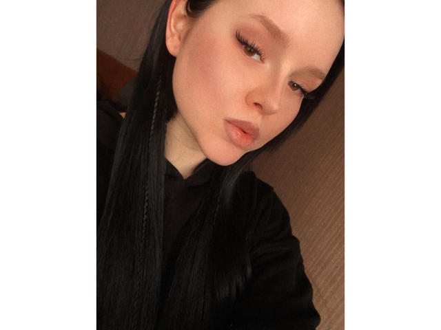 MillieLavinia has long black hair, pale skin, and pouty pink lips, wearing a black shirt. She looks slightly down with half-closed eyes and subtle eyeliner.