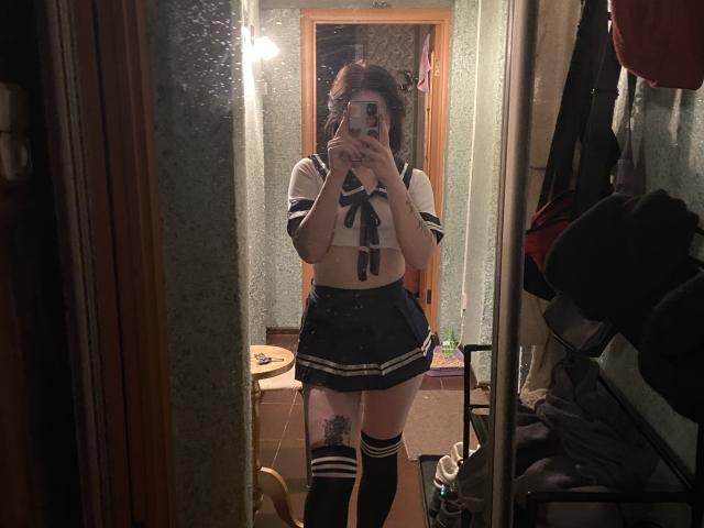 MillieLavinia in a mirror selfie wearing a navy and white schoolgirl outfit with a tied-up top, short skirt, and thigh-high socks. Visible tattoo on her right thigh.