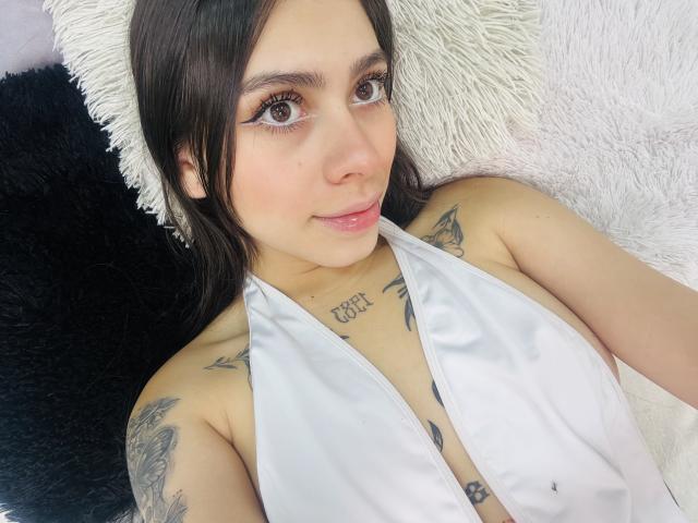 ChanelDeRosee, con cabello oscuro, un top blanco de tirantes, tatuajes en el pecho y los hombros, y delineador de ojos, acostada sobre almohadas blancas y negras, sonriendo a la cámara..