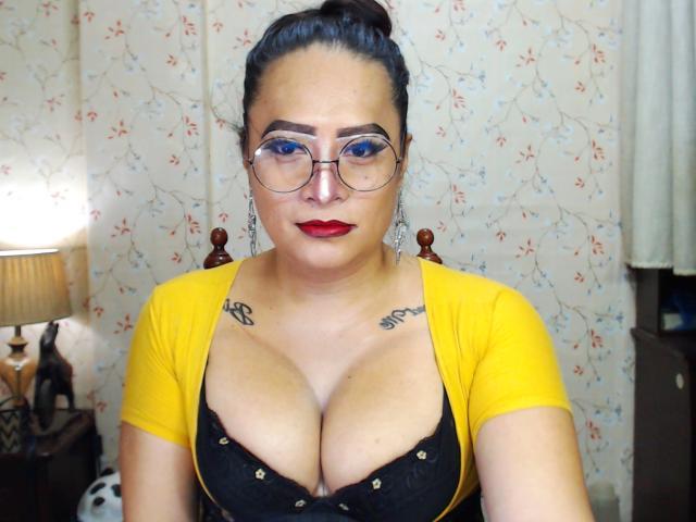 SquirtCumAlots with glasses, red lipstick, yellow top, black lace bra, large tits, tattoos on chest, dark hair in bun, standing in room, looking at camera, serious expression.