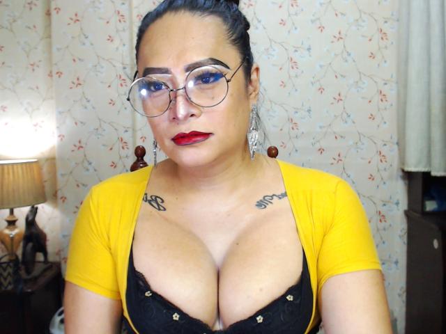 SquirtCumAlots with dark hair in a bun, wearing glasses, red lipstick, yellow cardigan, black lace bra, large tits, tattoos on chest, earrings, looking serious, in a bedroom.