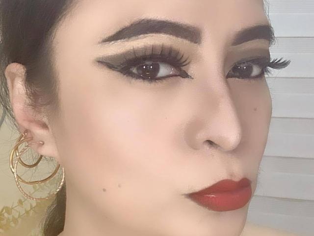 SquirtCumAlots has dark eyebrows, long black eyelashes, red lipstick, and gold hoop earrings, with a slightly serious face and brown eyes, in a sharp, well-lit selfie.