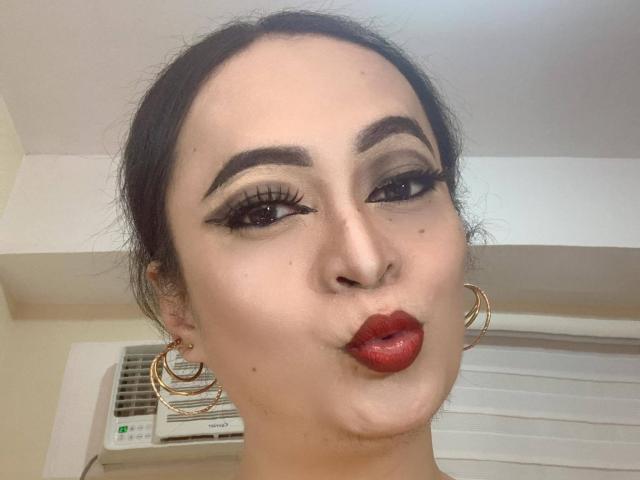 SquirtCumAlots has dark hair, thick eyebrows, long eyelashes, red lipstick, and gold hoop earrings, puckering their lips in a selfie. Their skin is fair and makeup is bold.