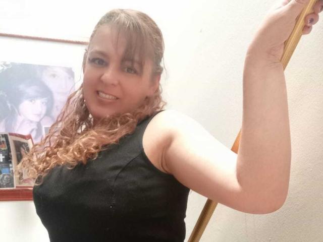 AmandaHotL with curly red hair, wearing a black sleeveless top, smiling, holding a wooden pole, showing her arm and upper body, tits slightly visible, in a photo.