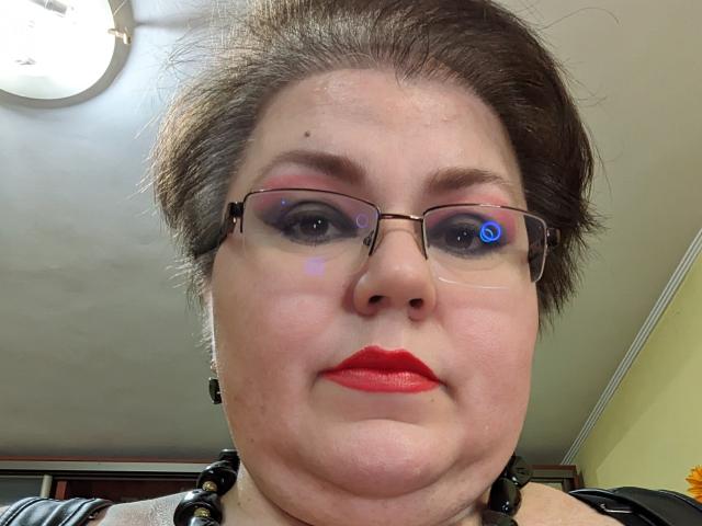 KaryQueen has short brown hair, glasses, red lipstick, and a slight double chin, with blue reflection in her eyes, wearing black beaded necklace, looking at the camera.