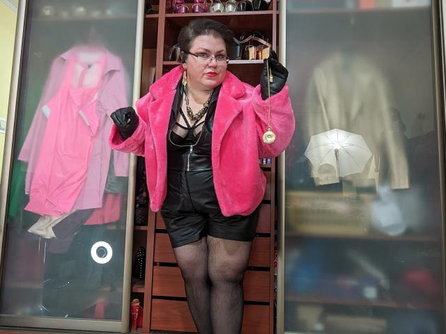 KaryQueen with pink fur jacket, black leather shorts, black stockings, and red lipstick, standing in front of a wooden closet with pink and beige jackets, holding a necklace.