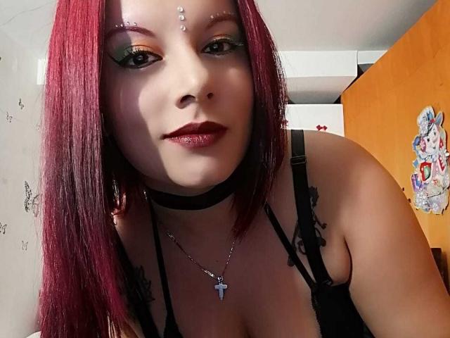 SexyAfroditaX with red hair, black choker, and cross necklace, wearing black lingerie, has tattoos on shoulders, green eye makeup, and red lipstick, looking at camera with a slight smile.