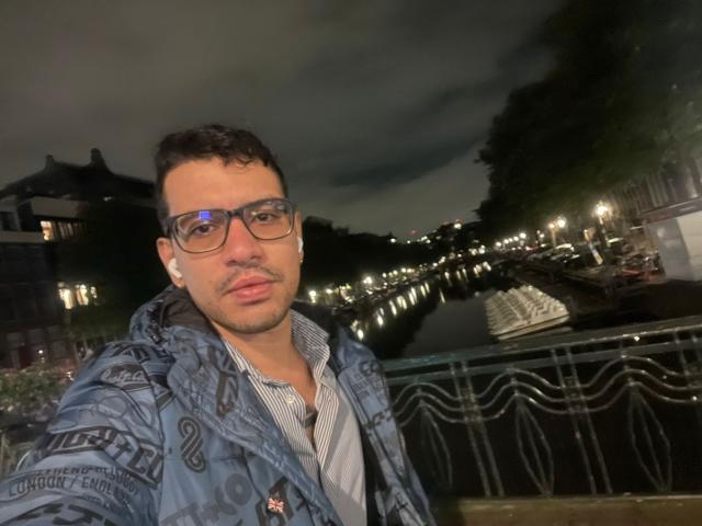 Damian Delatorre, portant des lunettes, avec des cheveux courts et bouclés bruns, peau claire, portant une veste bleue à motifs par-dessus une chemise rayée, se tient près d'un canal illuminé la nuit..