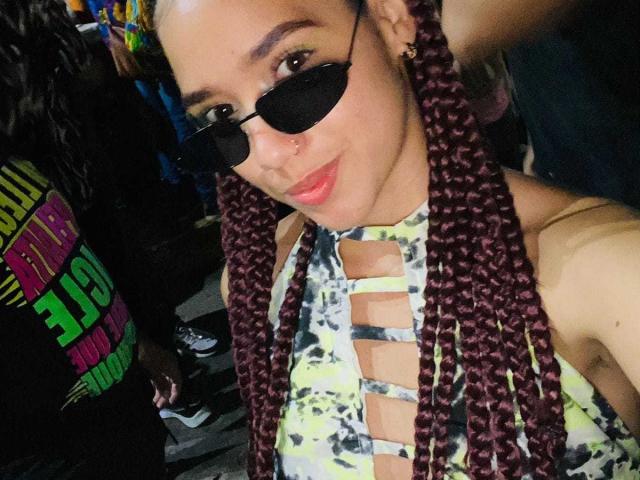 CandyCandyHot with purple braids, black sunglasses, nose ring, and a tie-dye top with cutout sections, smiling at the camera. Boobs visible through top's cutouts.