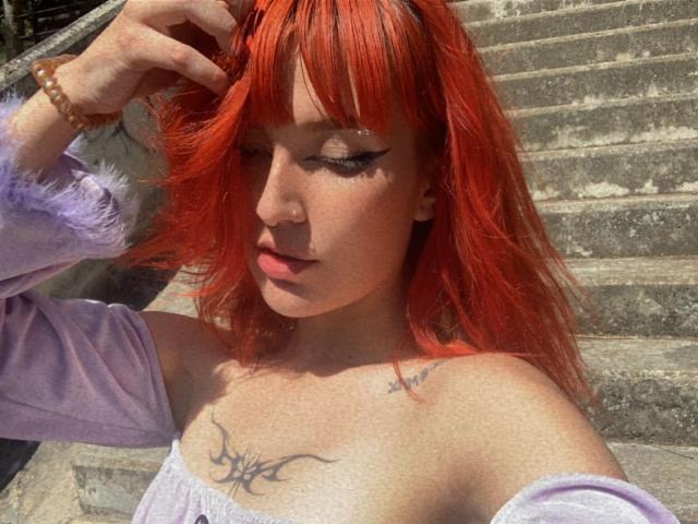 MaryWane with bright red hair, off-shoulder purple top, and black tribal tattoo on chest, stands on sunlit stairs, hand in hair, looking down, wearing a bead bracelet.
