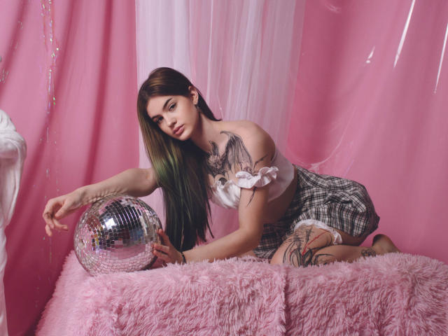 Nicole, mit langen, braunen Haaren mit grünen Spitzen, einem weißen Top mit einem Schulterauschnitt, einem karierten Rock und Tattoos, kniet auf einer rosa, flauschigen Decke, hält einen glänzenden, silbernen Ball und schaut in die Kamera..