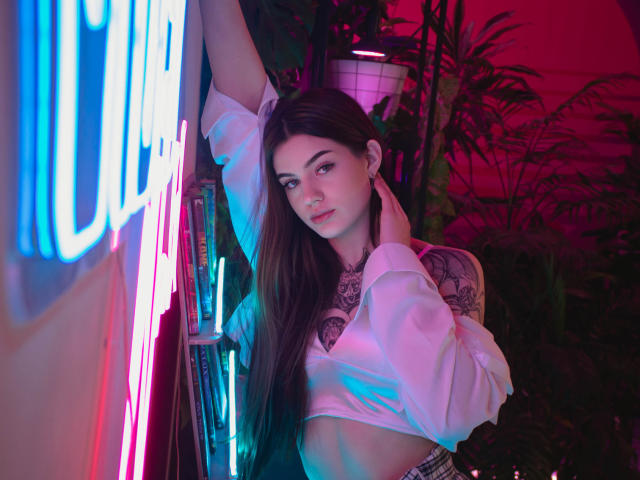 Nicole, avec de longs cheveux bruns, une chemise blanche à manches bouffantes, des tatouages sur la poitrine et l'épaule, appuyée contre un mur bleu néon et rose, une main sur le cou, éclairage rose et violet..